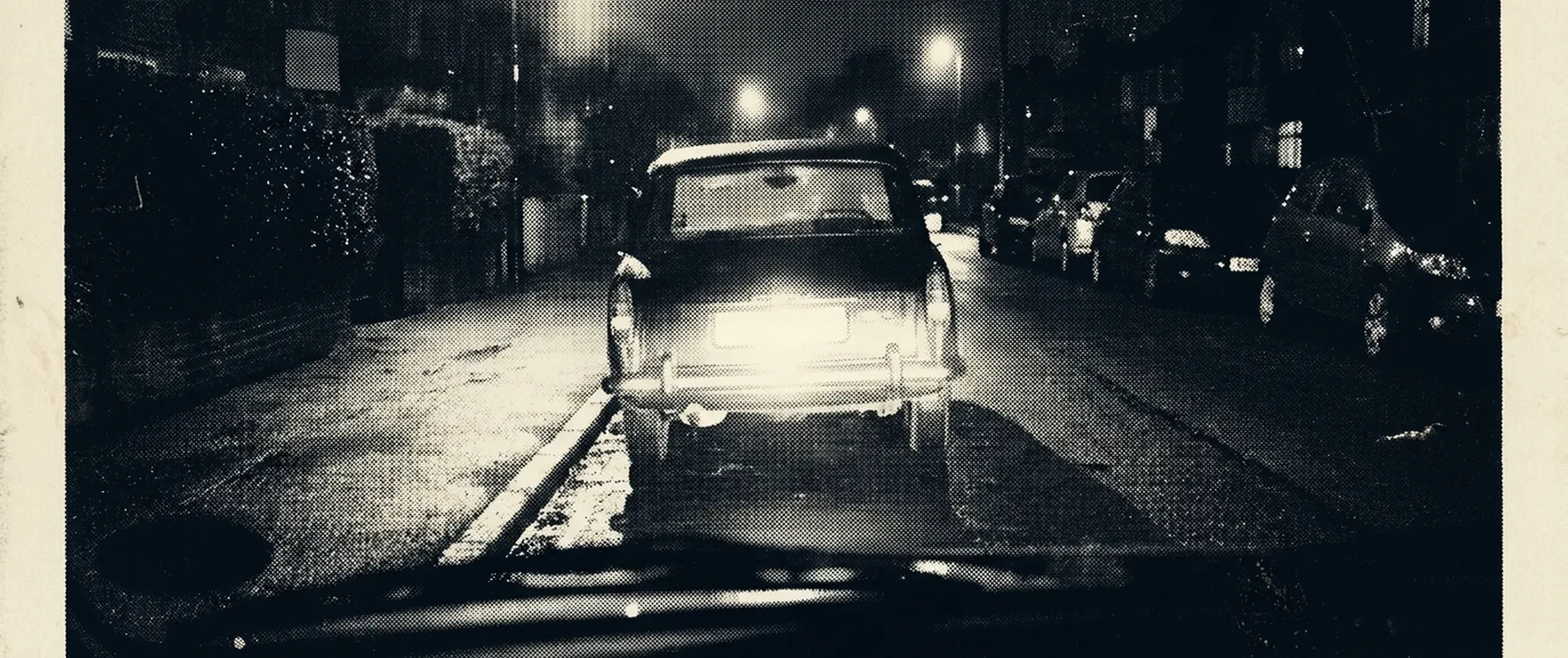 A dark suburban road at night seen from a windscreen, with the number plate of the car ahead blown out by headlight glare into an unreadable bright rectangle.